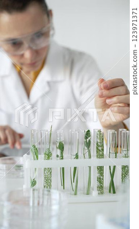 Woman scientist wearing a lab coat, is dripping liquid from a pipette into a test tube with a green plant inside, vertical close up view. Science and medicine concepts 123971371