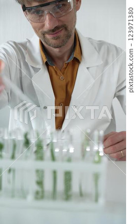Man scientist wearing a lab coat and protective glasses, is dripping liquid from a pipette into a test tube with a green plant inside, face focused vertical portrait. Science and medicine 123971380