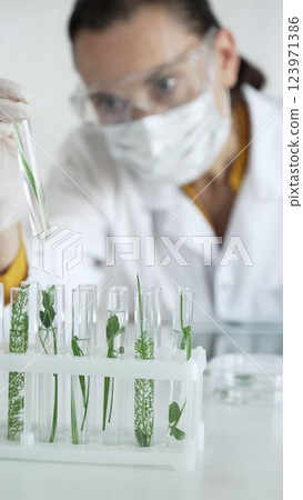 Woman scientist with white gloves, face mask and protective glasses, is holding a test tube with plants inside in laboratory setting, vertical view. Science and medicine Woman scientist with white gloves, face mask and protective glasses, is holding a test tube with plants inside in laboratory setting, vertical view. Science and medicine 123971386
