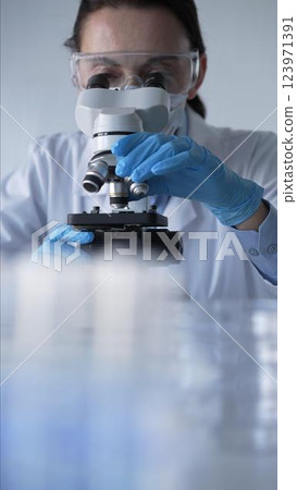 Woman scientist wearing blue gloves, protective glasses and medical mask is using microscope in modern laboratory setup. Low key lighting photo. Science and Medicine Woman scientist wearing blue gloves, protective glasses and medical mask is using microscope in modern laboratory setup. Low key lighting photo. Science and Medicine 123971391