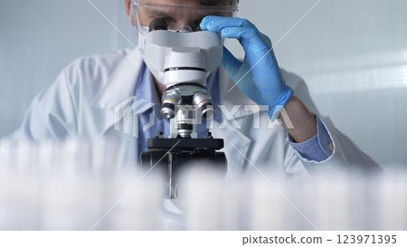 Male scientist researcher wearing blue gloves, protective glasses and medical mask, is using microscope in modern laboratory, portrait view. Low key lighting photo. Science and Medicine 123971395