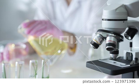A scientist, wearing a lab coat and pink protective gloves, is pouring a yellow oily liquid from one beaker to another near microscope in laboratory, close up. Medicine and science A scientist, wearing a lab coat and pink protective gloves, is pouring a yellow oily liquid from one beaker to another near microscope in laboratory, close up. Medicine and science 123971396