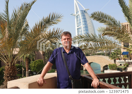A confident adult man poses for a photo against the stunning backdrop of a famous hotel in Dubai, showcasing the luxurious architecture and vibrant atmosphere of the city, capturing a moment of joy 123971475