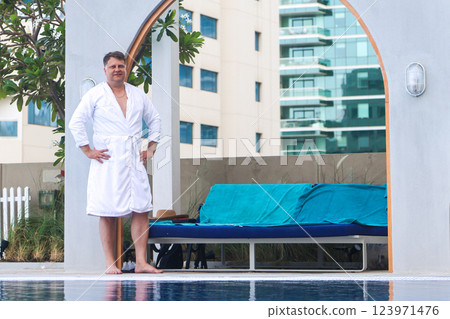 A Man in a White Robe Stands by the Hotel Pool, Preparing to Dive into the Refreshing Water, Embracing a Day of Leisure and Comfort Under the Bright Sun, Ready to Unwind and Enjoy His Vacation 123971476
