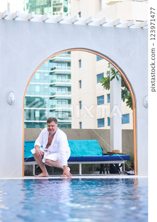 A Man in a White Robe Stands by the Hotel Pool, Preparing to Dive into the Refreshing Water, Embracing a Day of Leisure and Comfort Under the Bright Sun, Ready to Unwind and Enjoy His Vacation 123971477