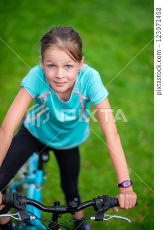 Smiling young girl riding a blue bicycle outdoors in a green park, enjoying an active lifestyle and outdoor fun in summer 123971498