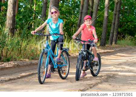 Two smiling young girls riding bicycles on a dirt road through a green forest on a sunny day, enjoying outdoor adventure and summer fun Two smiling young girls riding bicycles on a dirt road through a green forest on a sunny day, enjoying outdoor adventure and summer fun 123971501
