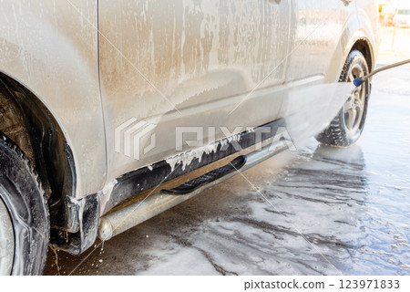 Close-up of a car being washed with high-pressure water and foam at a self-service car wash, focusing on the wheel and side panel covered in soap suds Close-up of a car being washed with high-pressure water and foam at a self-service car wash, focusing on the wheel and side panel covered in soap suds 123971833