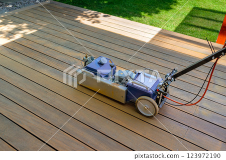 Worker cleaning and sanding a wooden deck with a specialized machine in a backyard setting with lush green lawn and a bright sunny environment. 123972190