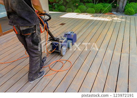 Worker cleaning and sanding a wooden deck with a specialized machine in a backyard setting with lush green lawn and a bright sunny environment. 123972191