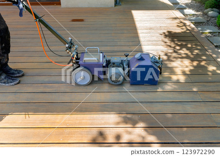 Worker cleaning and sanding a wooden deck with a specialized machine in a backyard setting with lush green lawn and a bright sunny environment. Worker cleaning and sanding a wooden deck with a specialized machine in a backyard setting with lush green lawn and a bright sunny environment. 123972290