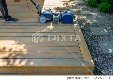 Worker cleaning and sanding a wooden deck with a specialized machine in a backyard setting with lush green lawn and a bright sunny environment. Worker cleaning and sanding a wooden deck with a specialized machine in a backyard setting with lush green lawn and a bright sunny environment. 123972291