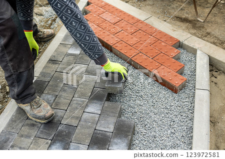 Construction worker is building a sidewalk, placing gray concrete paving stones on gravel 123972581