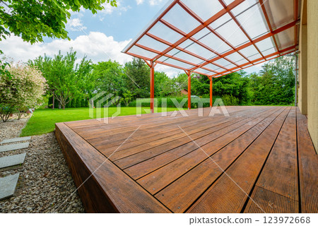 Wooden garden deck with a polycarbonate pergola roof, surrounded by a lush green backyard and landscaping, providing a shaded outdoor relaxation area 123972668