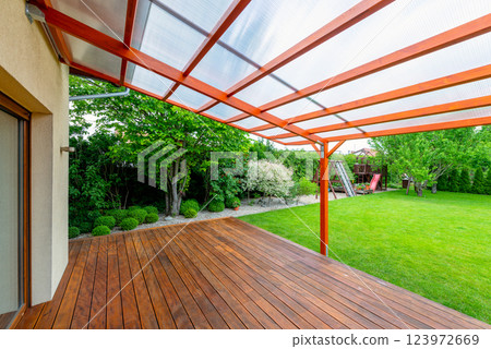 Wooden pergola with transparent polycarbonate roof covering a garden deck in a modern backyard, featuring a lush green lawn and landscaping for outdoor relaxation. 123972669