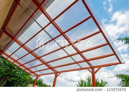 Transparent polycarbonate roof on a modern wooden pergola structure in a backyard, providing shade and protection with a clear sky background. Transparent polycarbonate roof on a modern wooden pergola structure in a backyard, providing shade and protection with a clear sky background. 123972670