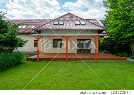 Modern suburban house with a red tiled roof, wooden deck with a transparent pergola, and a beautifully landscaped backyard featuring green grass and lush trees on a sunny day Modern suburban house with a red tiled roof, wooden deck with a transparent pergola, and a beautifully landscaped backyard featuring green grass and lush trees on a sunny day 123972674