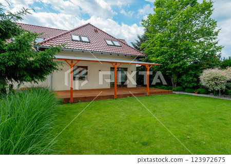 Modern suburban house with a red tiled roof, wooden deck with a transparent pergola, and a beautifully landscaped backyard featuring green grass and lush trees on a sunny day Modern suburban house with a red tiled roof, wooden deck with a transparent pergola, and a beautifully landscaped backyard featuring green grass and lush trees on a sunny day 123972675