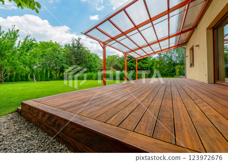 Wooden garden deck with a polycarbonate pergola roof, surrounded by a lush green backyard and landscaping, providing a shaded outdoor relaxation area 123972676