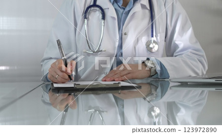 Woman doctor, wearing a blue blouse under a white medical coat, is using a clipboard and taking notes on glass desk in a medical office, close-up. Medicine 123972890