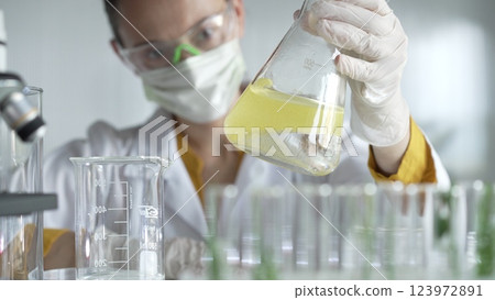Woman scientist researcher wearing white protective gloves is shaking up a yellow chemical solution inside an Erlenmeyer flask in a laboratory setting, portrait view. Science and medicine 123972891