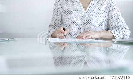 Businesswoman in white dotted blouse is signing a contract at a sleek glass desk in a modern office, showcasing professionalism and focus in her work environment. Business people concept 123972910