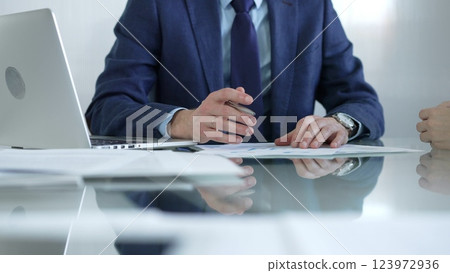 Businessman pointing at charts with pen, explaining data to colleague while sitting at the glass table in office. Businesspeople concept Businessman pointing at charts with pen, explaining data to colleague while sitting at the glass table in office. Businesspeople concept 123972936