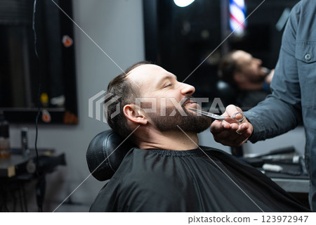 Skilled barber grooming clients beard using professional scissors, delivering precise styling within contemporary barbershop setting 123972947
