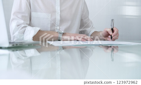 Businessman in white casual shirt is signing a contract at a sleek glass desk in a modern office, showcasing professionalism and focus in his work environment. Business people concept 123972962
