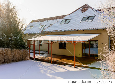 A cozy modern house with a snow-covered roof and wooden terrace under clear winter sunlight 123972965