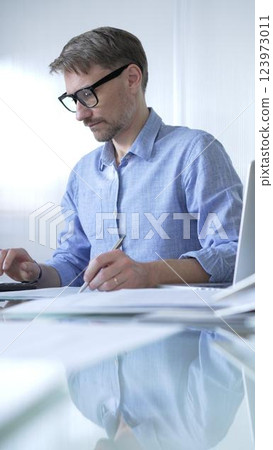 Businessman or accountant, wearing blue casual shirt and glasses, is working on tax calculations, note taking, and financial reports in the office. Audit and taxes in business 123973011