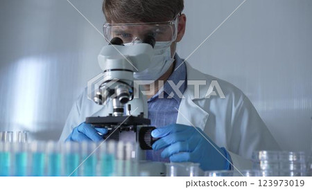 A scientist man, wearing a lab coat, mask, blue gloves and safety glasses, is using a binocular microscope surrounded by test tubes in laboratory. Portrait view. Medicine and science A scientist man, wearing a lab coat, mask, blue gloves and safety glasses, is using a binocular microscope surrounded by test tubes in laboratory. Portrait view. Medicine and science 123973019
