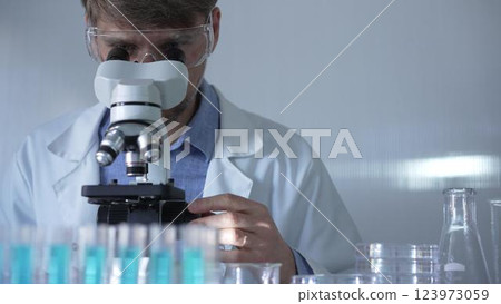A man scientist, wearing a lab coat, mask, and safety glasses, is using a microscope surrounded by test tubes in laboratory setting. Medicine and science concept A man scientist, wearing a lab coat, mask, and safety glasses, is using a microscope surrounded by test tubes in laboratory setting. Medicine and science concept 123973059