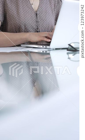Businesswoman, wearing brawn dotted blouse, is typing on laptop keyboard, preparing taxes and audit reports, with documents on glass office desk, vertical view. Taxes, audit in business 123973242