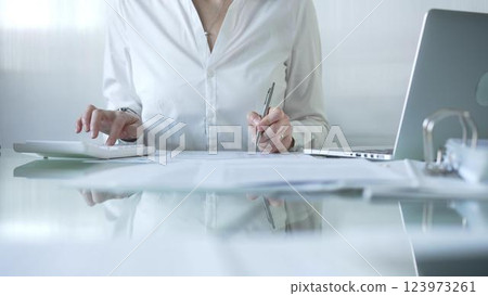 Businesswoman in blue blouse is using white calculator and taking notes while working on financial report in contemporary office setting. Audit and taxes concept 123973261