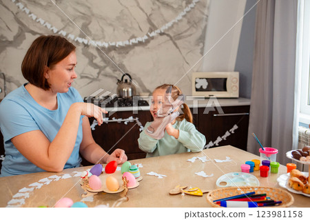 Smiling mother and cute little girl play together with rabbit glove, sitting at Easter table. Spring holiday Smiling mother and cute little girl play together with rabbit glove, sitting at Easter table. Spring holiday 123981158