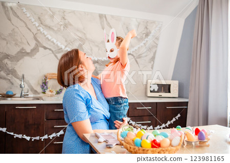 Wide shot of Caucasian happy mother and her child are hugging nearby festive table for Easter. A cute little girl in a paper rabbit mask Wide shot of Caucasian happy mother and her child are hugging nearby festive table for Easter. A cute little girl in a paper rabbit mask 123981165