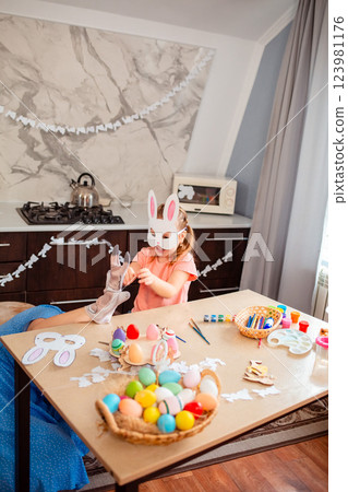 Wide shot of young mother plays with rabbit glove with her little daughter in a decorated kitchen. Funny girl in a paper rabbit mask is having fun with her mother. Vertical 123981176