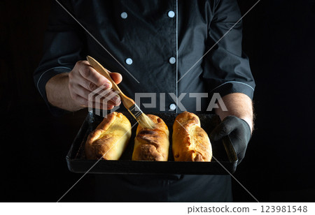 A skilled chef holding a tray of three golden rolls applies a glossy glaze with a brush. The kitchen background is dimly lit, emphasizing the texture of the bread A skilled chef holding a tray of three golden rolls applies a glossy glaze with a brush. The kitchen background is dimly lit, emphasizing the texture of the bread 123981548