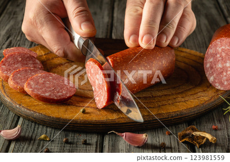 A person skillfully slices a sausage on a rustic wooden board. The kitchen has a warm atmosphere, highlighting the art of gourmet food preparation with fresh ingredients A person skillfully slices a sausage on a rustic wooden board. The kitchen has a warm atmosphere, highlighting the art of gourmet food preparation with fresh ingredients 123981559