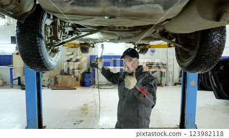 Confident automotive technician smiling, giving thumbs up, holding inspection lamp under lifted vehicle at modern garage workshop 123982118