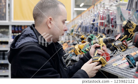 Concentrated male customer choosing cordless drill in hardware store, examining battery and chuck type, comparing power tools, making purchase decision in diy shop Concentrated male customer choosing cordless drill in hardware store, examining battery and chuck type, comparing power tools, making purchase decision in diy shop 123982122