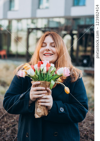 Beautiful young woman with spring tulips flowers bouquet walking at city street. Happy girl posing outdoors. Spring portrait of pretty female on urban background. High quality photo. 123982414