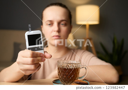 A person is checking their blood sugar levels using a glucometer while sitting at a table with a cup of tea and cookies. Soft lighting creates a warm atmosphere A person is checking their blood sugar levels using a glucometer while sitting at a table with a cup of tea and cookies. Soft lighting creates a warm atmosphere 123982447