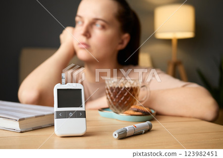 A woman sits thoughtfully at a table with a glucometer, a cup of tea, and snacks. She appears contemplative, focusing on managing her health during a serene afternoon A woman sits thoughtfully at a table with a glucometer, a cup of tea, and snacks. She appears contemplative, focusing on managing her health during a serene afternoon 123982451