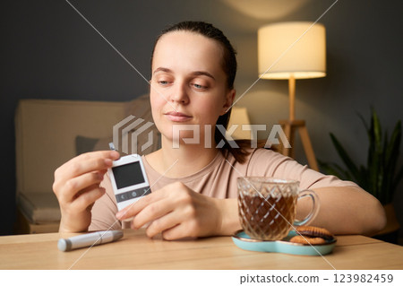 A woman sits at a table in a cozy room examining her glucometer. She has a warm beverage beside her and a plate of cookies, reflecting a calm evening routine focused on health A woman sits at a table in a cozy room examining her glucometer. She has a warm beverage beside her and a plate of cookies, reflecting a calm evening routine focused on health 123982459