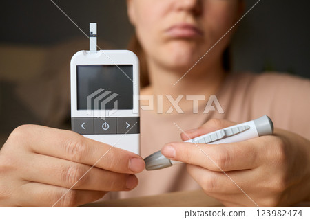 A person holds a glucometer and a pen, appearing worried while preparing to check blood sugar levels at home during the day. The setting is calm and focused on health management 123982474