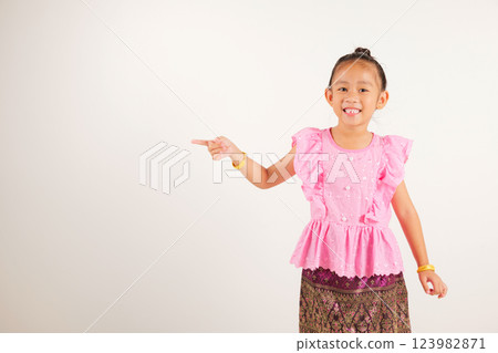 Portrait Thailand kid girl smiling traditional Thai dress costume indicate finger empty space, studio isolated white background, kindergarten child pointing index out with copy space 123982871