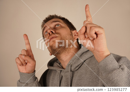 A young man with dark hair points upward with both index fingers, emphasizing something significant. His focused expression adds meaning to the gesture. The pose conveys importance and attention. A young man with dark hair points upward with both index fingers, emphasizing something significant. His focused expression adds meaning to the gesture. The pose conveys importance and attention. 123985277