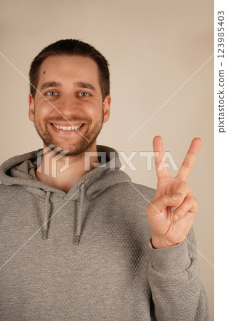 A young man in a grey hoodie makes the peace sign with his fingers, forming a "V" shape. His casual gesture conveys a sense of harmony and positivity. The moment reflects peace and unity. 123985403